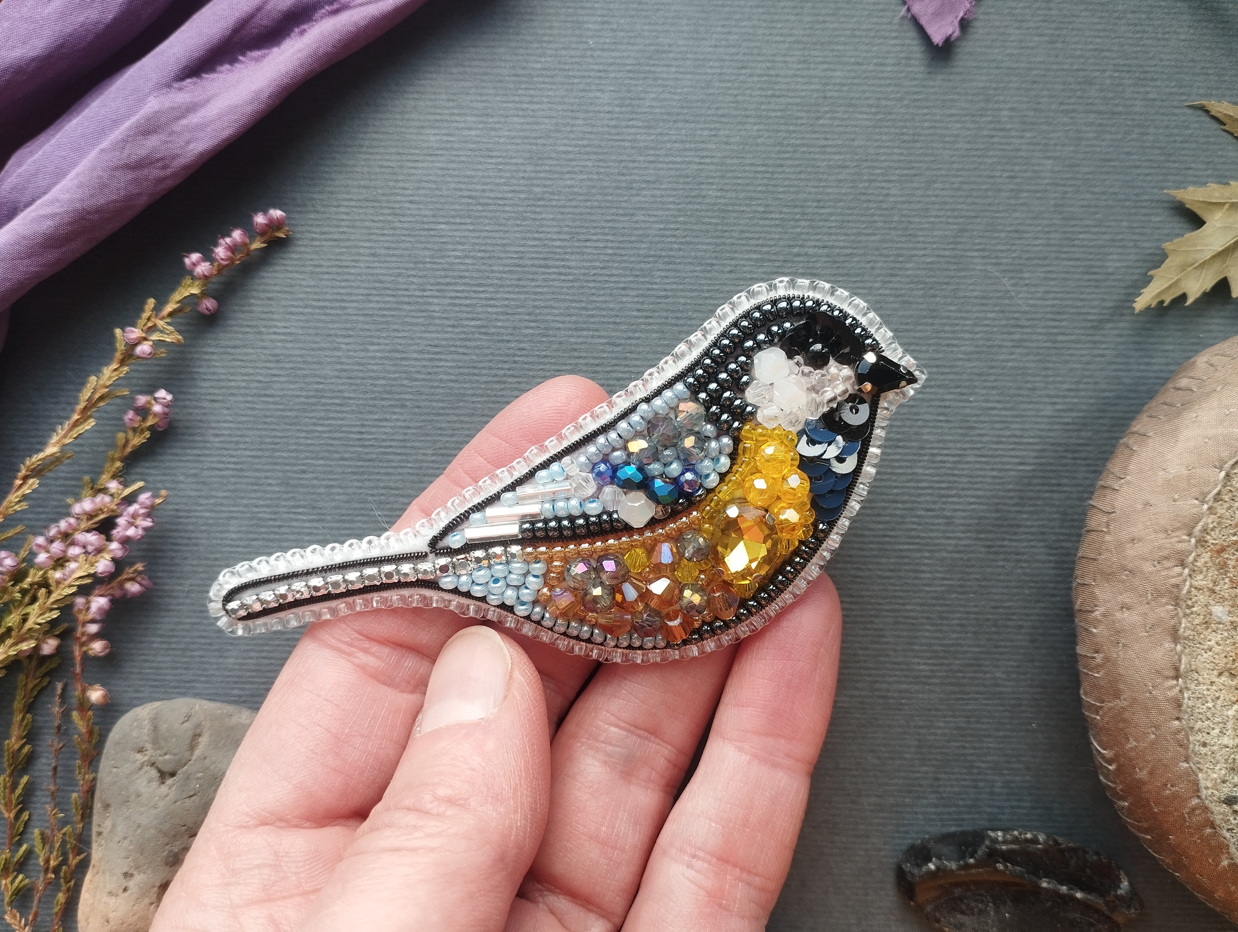 A hand is holding a decorative bird brooch made of colorful beads and sequins, with a purple fabric and some dried flowers in the background.